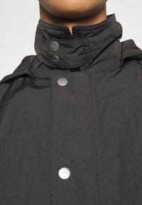 Giacca leggera nera con colletto alto. Presenta bottoni automatici, cuciture visibili e una texture stropicciata. Design minimalista.