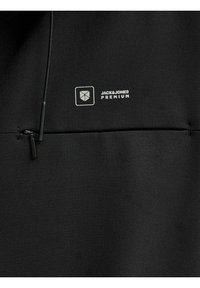 Material negru cu un buzunar cu fermoar, șnur și logo alb „Jack & Jones Premium” lângă o pictogramă geometrică de scut.