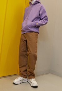 Felpa con cappuccio colore lavanda, dotata di tasca frontale, abbinata a pantaloni cargo color beige con tasche e cuciture gialle. Le sneaker bianche completano il look.