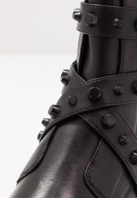 Botín de piel negra hasta el tobillo con acentos de tachuelas y correas en cruz. Textura suave con un acabado elegante y contrastes en hardware mate.