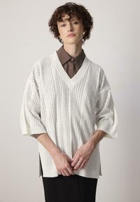 Pull en tricot blanc avec un col en V et des manches courtes et larges. Présente des lignes verticales et des détails perforés. Porté par-dessus une chemise marron à col.