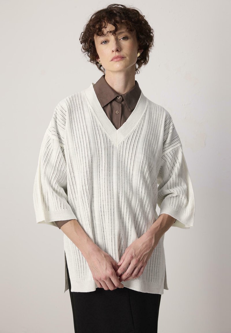 Pull en tricot blanc avec un col en V et des manches courtes et larges. Présente des lignes verticales et des détails perforés. Porté par-dessus une chemise marron à col.