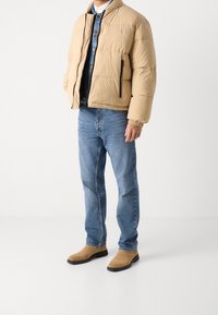 Beige puffatakki, jossa on pystysuora kaulus, vetoketjulla varustettu etuosa ja sivutaskut. Yhdistettynä sinisiin farkkuihin ja vaaleisiin nilkkasaappaisiin.