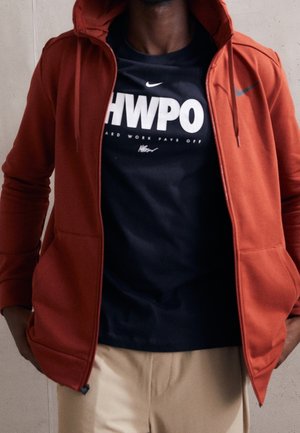 Red zip-up hoodie with a hood, featuring front pockets. Worn over a black T-shirt with white text, paired with light-colored pants.