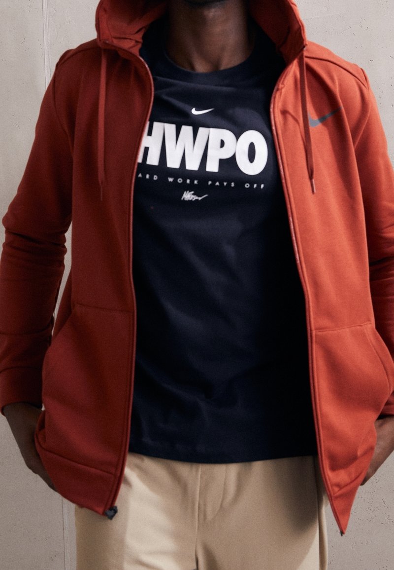 Red zip-up hoodie with a hood, featuring front pockets. Worn over a black T-shirt with white text, paired with light-colored pants.