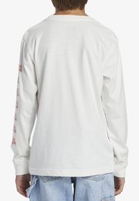 Weißes Langarm-T-Shirt mit Rundhalsausschnitt und buntem Textdesign, das entlang des linken Ärmels gedruckt ist, kombiniert mit hellblauen Denim-Shorts.