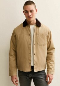 Ung man som bär en beige jacka med mörkrandig krage över en vit skjorta, stående mot en enkel ljus bakgrund.