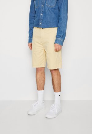 Shorts en coton jaune clair avec poches avant, associés à une chemise en denim bleu, des chaussettes blanches et des baskets blanches. Coupe décontractée et longueur mi-cuisse.