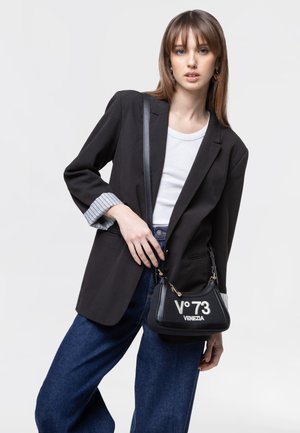 Giovane donna che indossa un blazer nero, un top bianco, jeans blu, tenendo una borsa nera con la scritta "V° 73 Venezia" su sfondo chiaro.