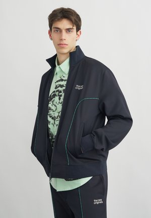 Fiatal férfi fekete cipzáras dzsekiben és hozzáillő, zöld szegélyes nadrágban, világoszöld mintás inggel, egyszínű háttér előtt áll.