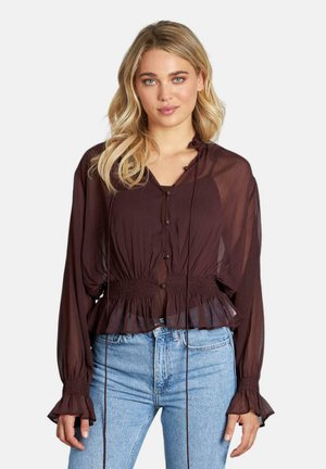 Blusa marrón transparente con volantes en el bajo y en los puños, con cierre frontal de botones y ajuste con cordón en la cintura. Combinada con jeans azules.