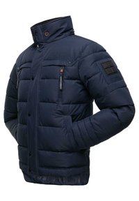 Chaqueta acolchada azul marino con cuello alto, bolsillos frontales con cremallera y un parche en la manga. Presenta una textura acolchada y cierre de botón.