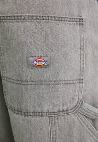 Gray denim apron featuring a front pocket with a colorful "Dickies" logo patch, reinforced stitching, and a tied waist strap for adjustment.