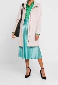 Persona che indossa un cappotto beige chiaro su una blusa verde e una gonna di raso teal, mentre porta una borsa tote nera e ha ai piedi un paio di décolleté neri con cinturino alla caviglia.