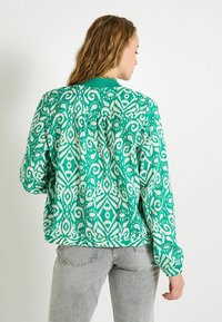 Chaqueta bomber verde con un diseño abstracto blanco, de mangas largas y con cuello de canalé. Llevada sobre unos vaqueros gris claro.