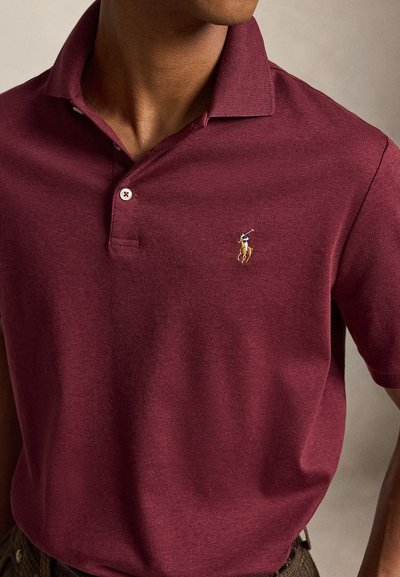 Inimese lähivõte, kes kannab tume-punast polot särki, mille vasakul rinnal on väike tikitud polo mängija hobusel logo.