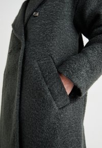 Abrigo de lana gris oscuro con una superficie texturizada, que presenta un cuello con solapa, dos botones y un bolsillo lateral. Diseño cálido y estructurado.