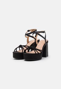 Sandali con tacco alto in suede nero con suola a piatto, caratterizzati da cinturini incrociati e un dettaglio annodato nella parte anteriore. Design con tacco a blocco.