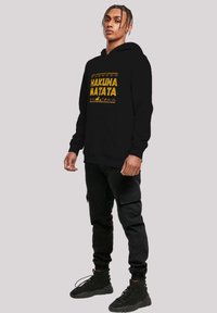 Fekete kapucnis pulóver, sárga színű "HAKUNA MATATA" felirattal és geometrikus mintákkal. Fekete cargo nadrággal és fekete lábbelivel párosítva.