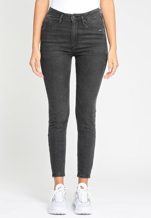 Mujer con vaqueros ajustados negros lavados y zapatillas blancas, de pie contra un fondo gris claro liso.