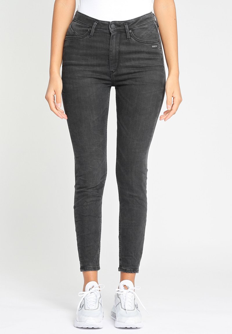 Nainen pukeutuneena istuviin mustiin kulutettuihin farkkuihin ja valkoisiin tennareihin, seisoo yksinkertaisen vaaleanharmaan taustan edessä.