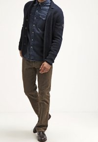 Mand iført en mørkeblå denimskjorte, navyblå cardigan, brune kordfløjlsbukser og mørkebrune sko, der står mod en hvid baggrund.