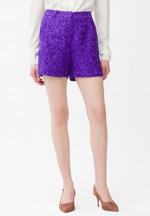 Shorts jacquard florales morados combinados con una camisa blanca de manga larga y zapatos de tacón alto marrones tejidos, en una mujer de pie.