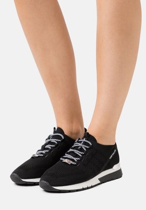 Zapatillas negras de punto con cordones blancos con estampado, usadas sobre pies desnudos, con suelas blancas y negras y un logo de "Tom Tailor" en el lateral.