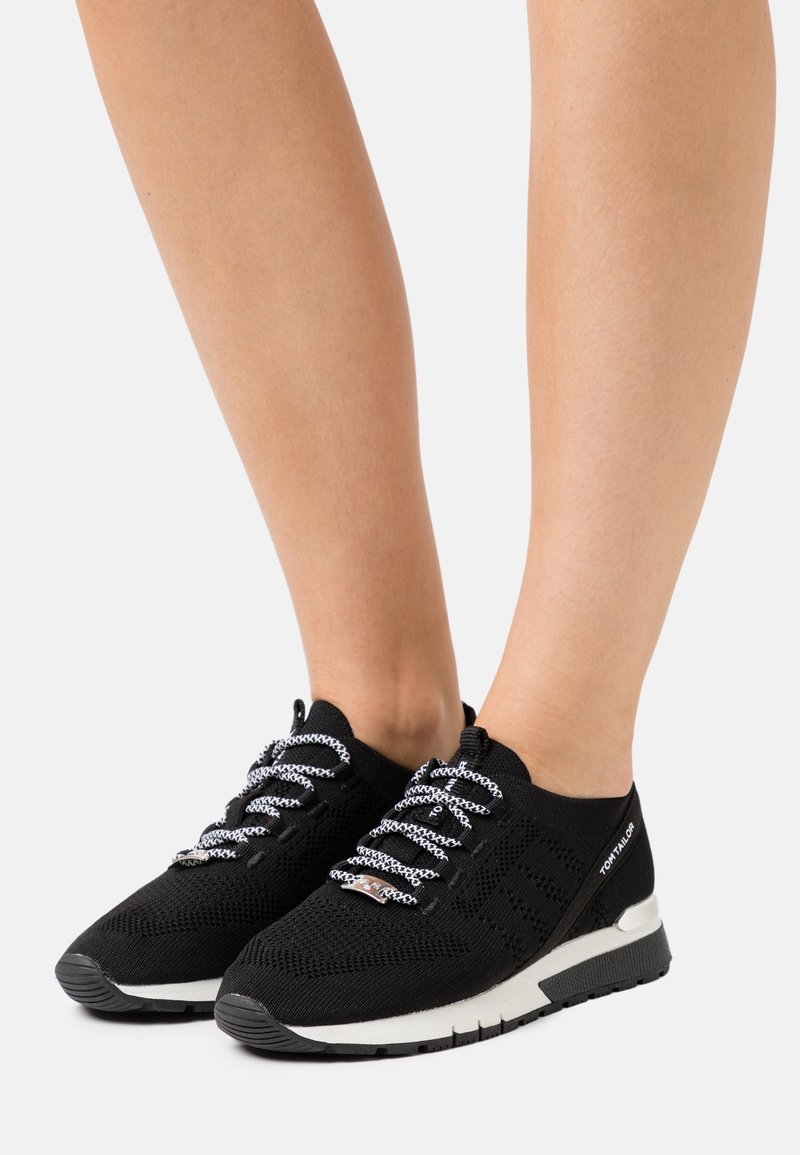 Czarne dzianinowe trampki z białymi sznurowadłami w wzory, noszone na bosych stopach, z białą i czarną podeszwą oraz logo "Tom Tailor" z boku.