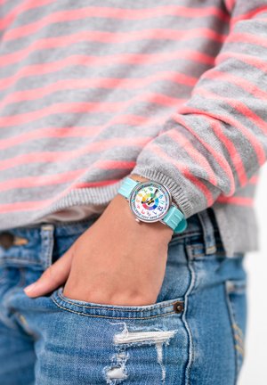 Montre colorée avec un boîtier rond en argent, un cadran blanc featuring des chiffres en gras, et un bracelet en silicone bleu clair, portée sur un pull rayé gris.