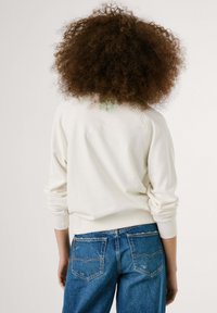 Witte sweatshirt met lange mouwen en een ronde hals, voorzien van een klein groen geborduurd patroon op de achterkant, gecombineerd met blauwe denim jeans.