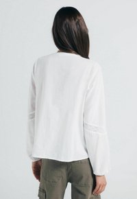 Hvid langærmet bluse med blonde accenter på ærmerne, afslappet pasform, let stof, let afrundet kant, parret med olivengrønne bukser.