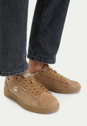 Sneakers G-Star in camoscio color cuoio con lacci, indossate con jeans neri, mostrano il logo laterale e cuciture dettagliate su uno sfondo chiaro e uniforme.