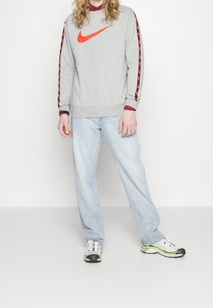 Persona che indossa una felpa grigia Nike con logo e strisce rosse, jeans azzurri, sneakers bianche con dettagli neri e verde neon.