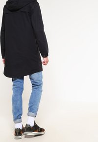Cappotto nero con cappuccio e retro lungo, abbinato a jeans a sigaretta blu chiaro e sneakers nere con dettagli arancioni. Calzini bianchi visibili.