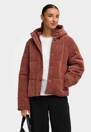 Femme portant une doudoune en velours côtelé couleur rouille avec capuche, chemise blanche, pantalon noir, tenant un petit sac noir dans une main.