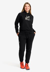 Svart hoodie med framficka, RVRC-logotyp i vitt, tillsammans med matchande svarta joggers. Klara orangea träningsskor ger en färgklick.