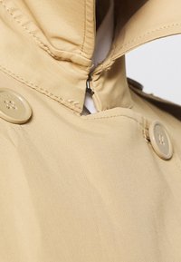 Impermeable beige con cuello alto, cierre frontal con cremallera y dos grandes botones redondos. Tela lisa y texturizada con detalles sutiles de costura.