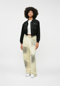 Črn skrčen jopič z narezanimi žepi, v kombinaciji s bež širokimi hlačami z temnimi madeži. Manekenka nosi belo majico in teniske.