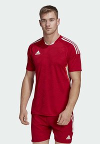 Jersey atletico rosso con motivo esagonale testurizzato, strisce bianche di accento sulle spalle e logo Adidas sul petto.