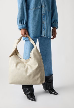 Cream woven tote bag with a slouchy shape, featuring a prominent seam and a small gold accent. Paired with a denim outfit and black boots.