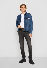 Chaqueta de denim con un lavado azul, camiseta blanca, jeans negros y zapatos negros. Presenta bolsillos frontales y un cinturón de marca.