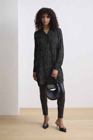 Zwart-witte blouse met stippen en kraag, gecombineerd met strakke zwarte leren broek en zwarte hoge hakken mules. Accessoire: kleine zwarte handtas.