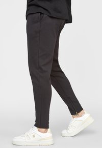 Mustat urheiluhousut kapealla leikkauksella, valmistettu pehmeästä materiaalista, joissa on sivutaskut ja nilkan kohdalla vetoketju. Valkoiset lenkkarit jalassa.