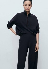 Femme portant un haut noir zippé avec les manches retroussées et un pantalon large assorti, devant un fond clair.
