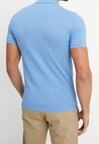 Ljusblå polo med slim fit, korta ärmar, ribbat krage och sidospår, visad på en person som står med ryggen mot kameran.