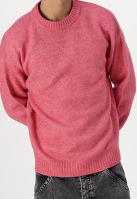 Pink knitted sweater with a ribbed crew neckline and cuffs, featuring a soft texture and slightly loose fit, paired with dark denim jeans.