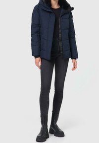 Granatowa kurtka puchowa z kapturem wykończonym futrem, pikowany wzór, zamek błyskawiczny z przodu oraz napy, zestawiona z czarnymi obcisłymi dżinsami i masywnymi czarnymi butami.