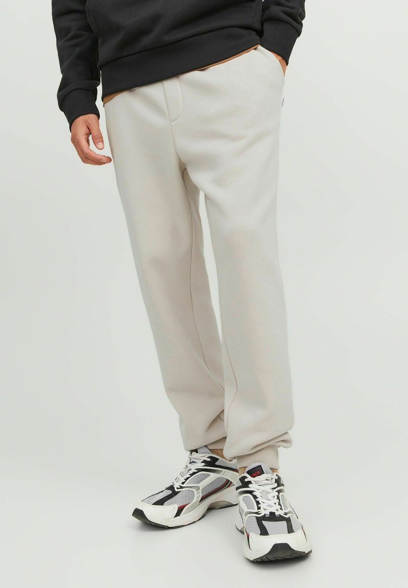 Hellgraue Jogginghose mit lockerem Schnitt, ausgestattet mit Seitentaschen und einer glatten Textur, kombiniert mit schwarz-weißen Sportschuhen.