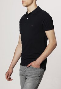 Granatowa koszulka polo z bawełny z klasycznym kołnierzykiem, zapinana na trzy guziki, z małym logo na piersi. Nosi się ją z jasnoszarymi dżinsami.